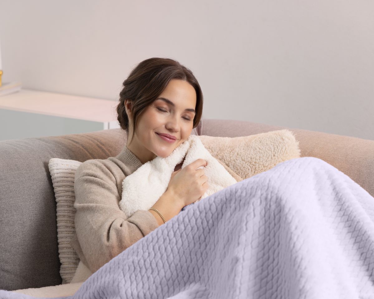 Woman cozily wrapped in a purple medisana heated blanket while relaxing on the sofa.